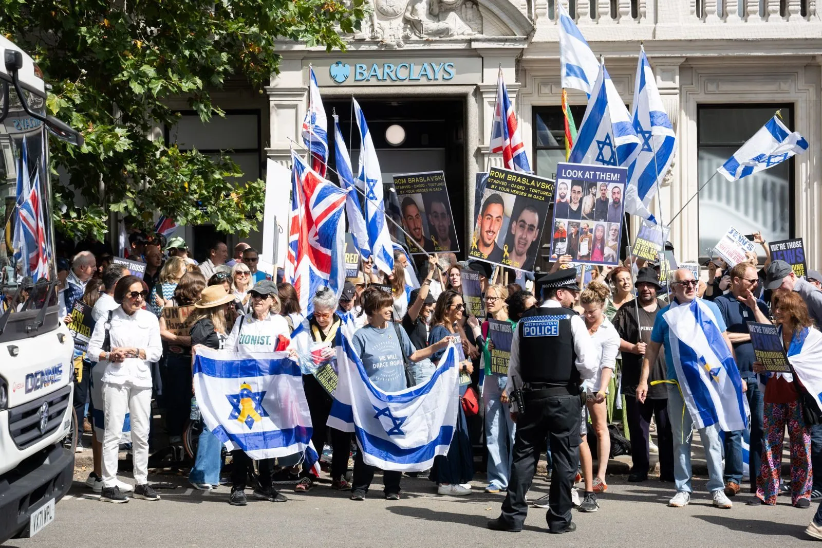 A police officer stands with his back to the camera in front of a demonstration outside a Barclays branch, Israeli flags and printed hostage posters behind him.