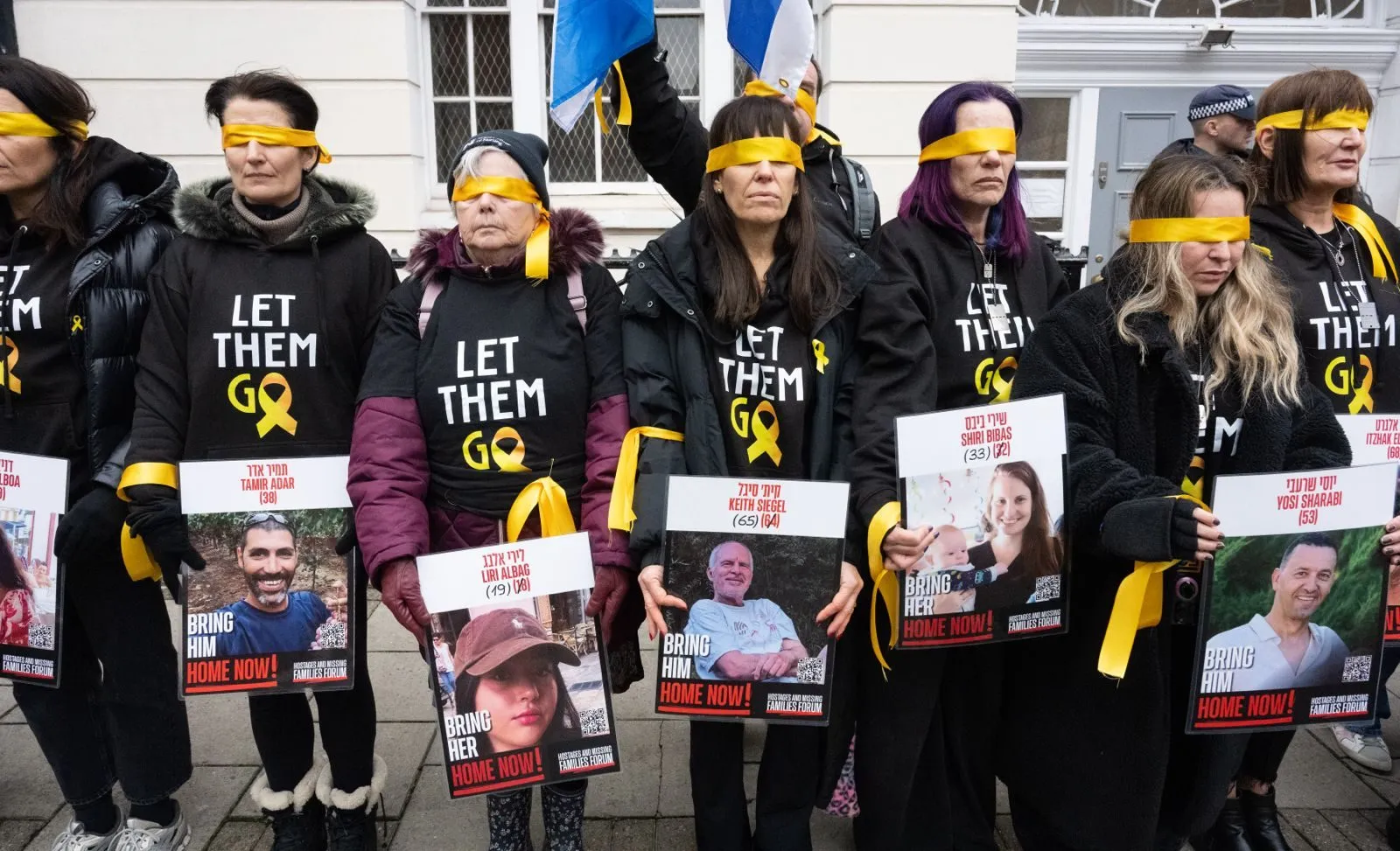 A row of people in Let Them Go shirts stand with yellow cloth tied across their eyes, each holding a printed poster of a named hostage.