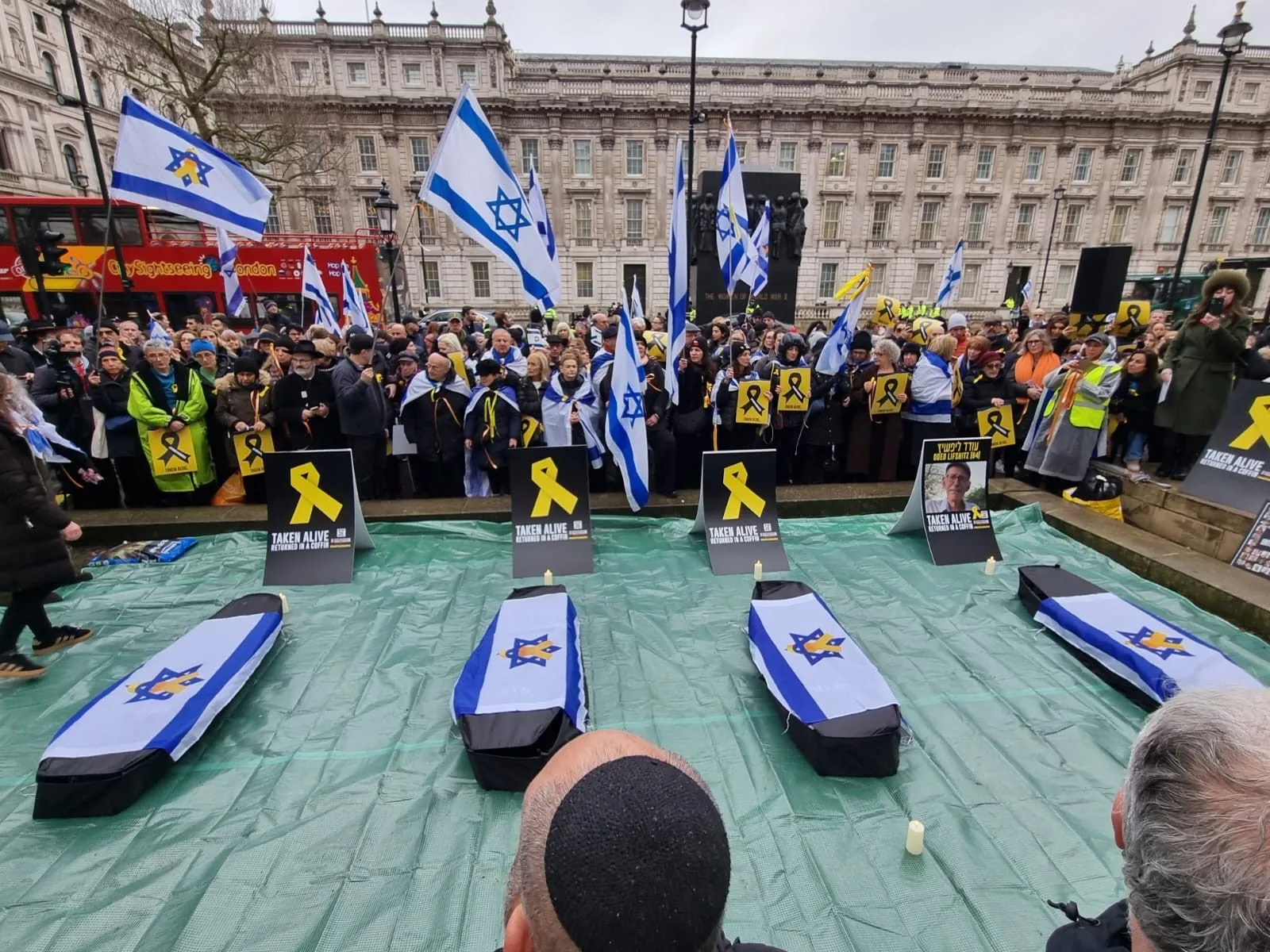 Four coffins draped in Israeli flags are laid on green sheeting in front of a crowd in Whitehall, each with a yellow ribbon placard beside it.