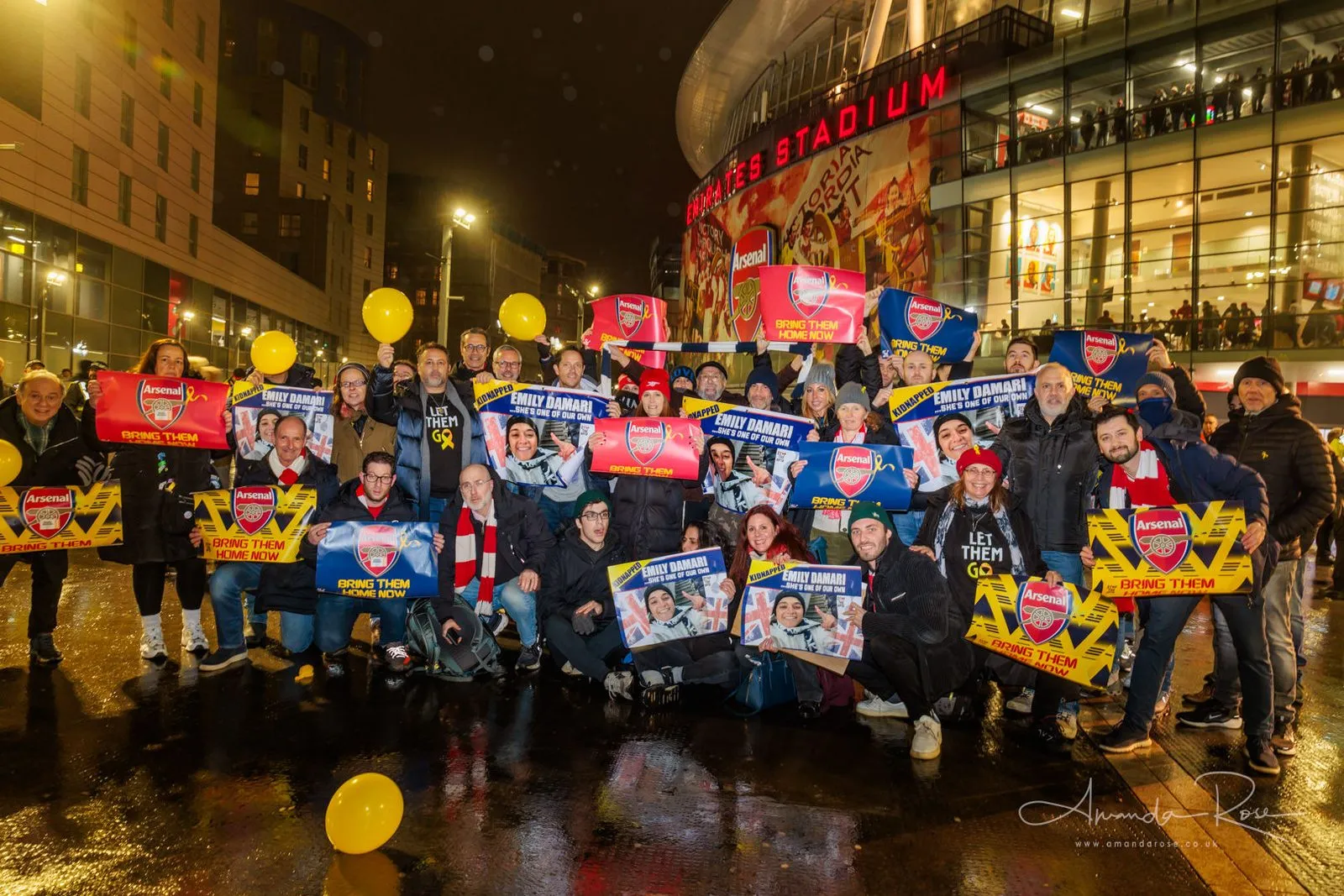 Arsenal and Tottenham supporters stand together in the rain outside the Emirates Stadium at night holding club scarves and hostage campaign placards.