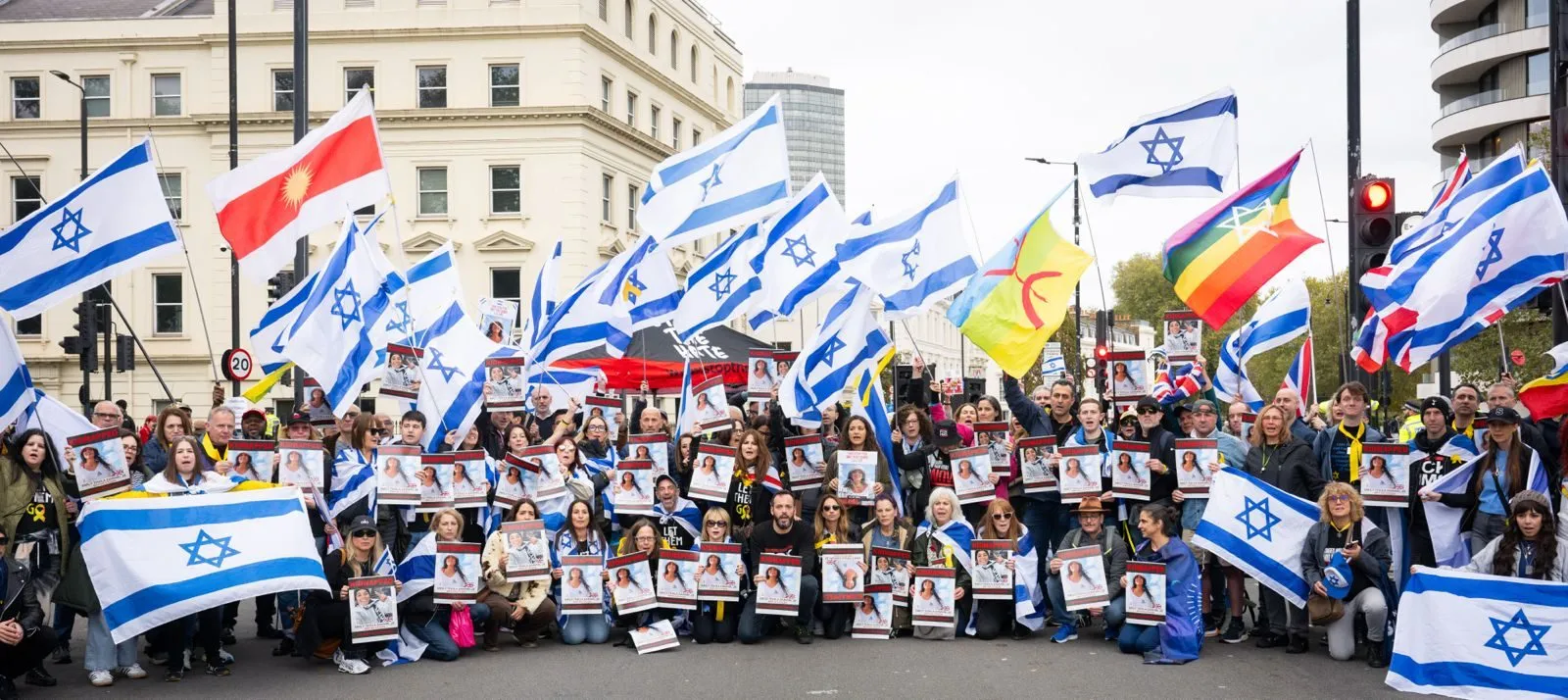 A wide group photograph of demonstrators filling a London street with Israeli and Union flags, many holding printed hostage posters at chest height.