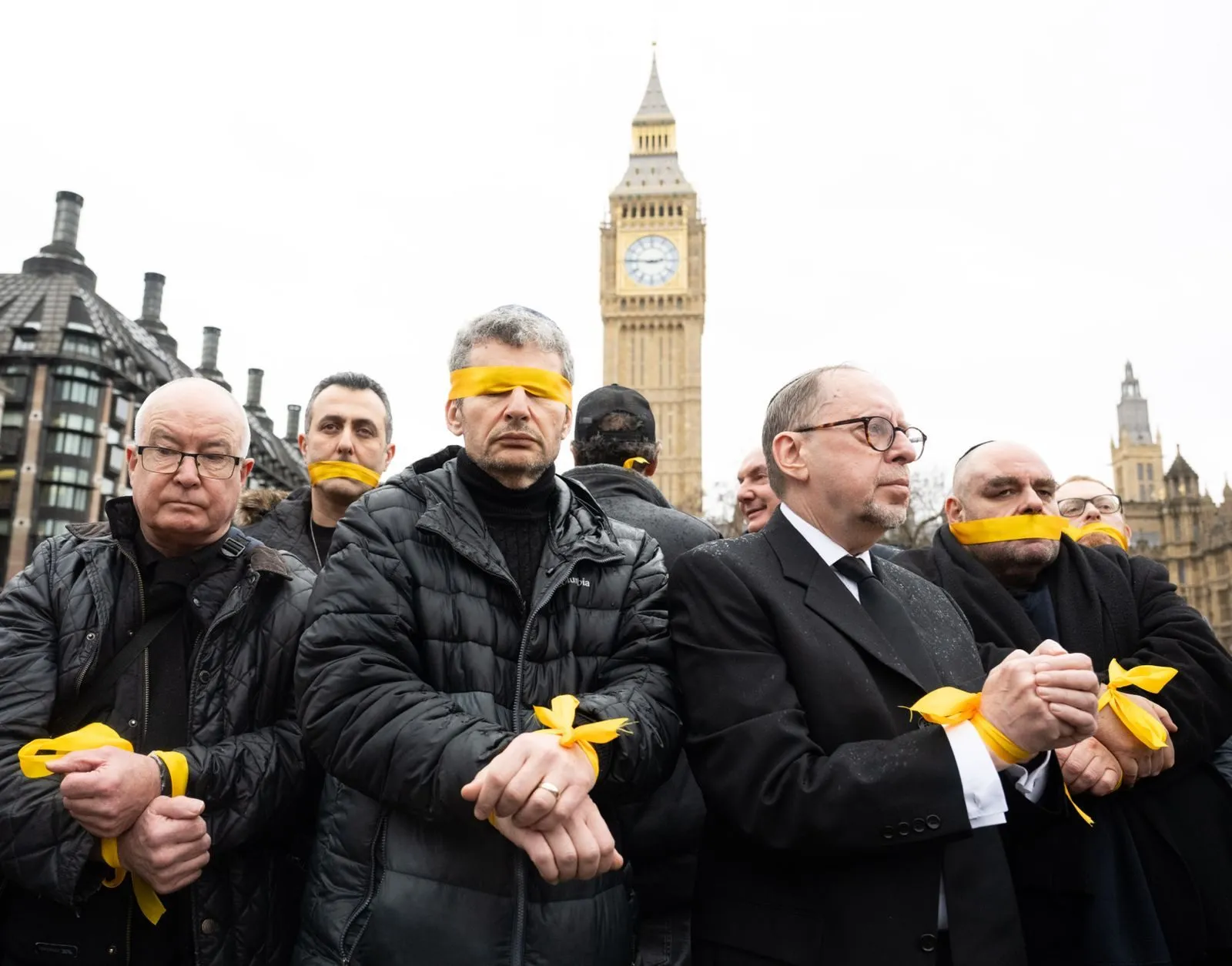 A row of men stand with wrists bound and yellow cloth tied over their mouths and eyes, the Elizabeth Tower behind them under a white sky.
