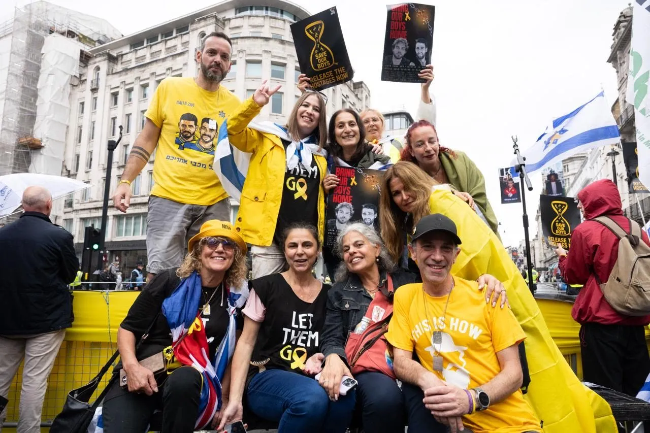 A group poses for a photograph in yellow shirts and hostage campaign badges at a road junction, holding printed posters above their heads.