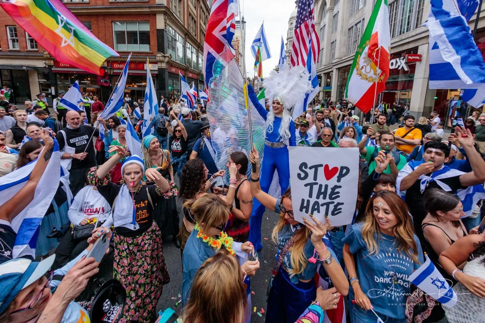 A woman holds a hand lettered Stop The Hate sign in a street parade, a performer on stilts in blue and white costume above the crowd behind her.