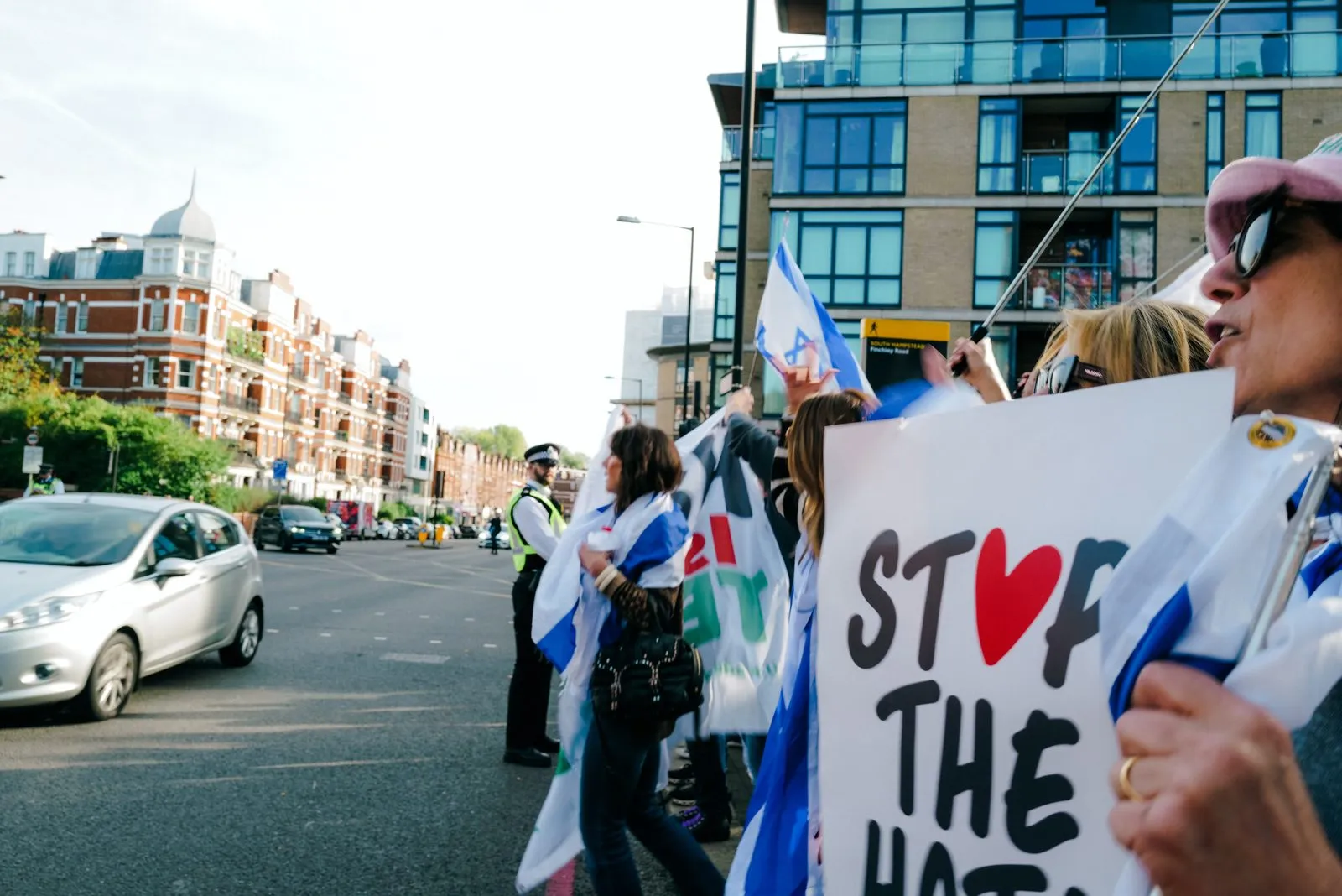 Hands hold a hand painted Stop The Hate placard at the edge of a road, a police officer and a line of demonstrators with Israeli flags further down the street.