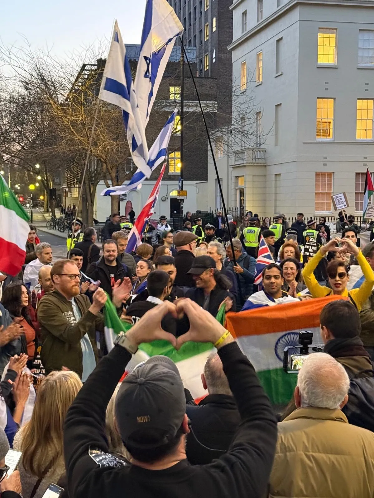 Several people in a crowd raise their hands to make heart shapes at dusk, Israeli flags and a lit Georgian terrace behind them.