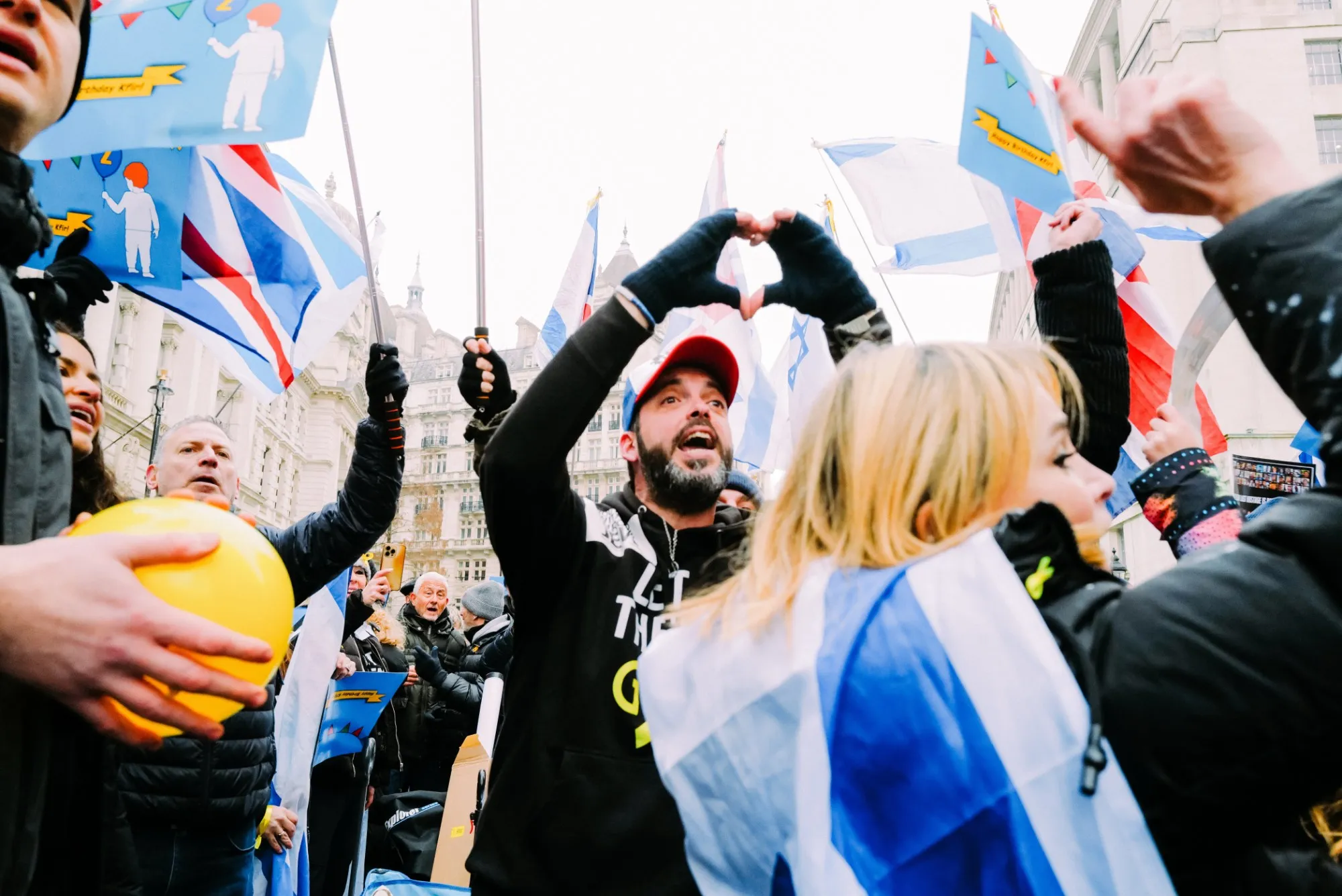 A man in a Let Them Go hoodie and a red cap raises both hands into a heart above his head in a winter crowd, Israeli and Union flags and blue Happy Birthday Kfir placards around him.