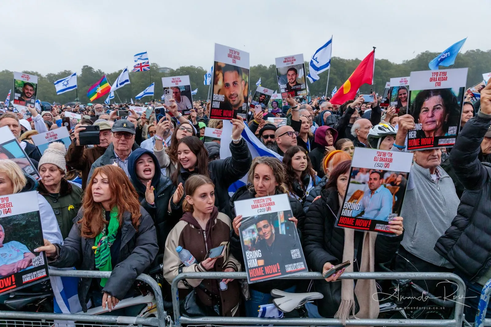Thousands of people fill Hyde Park in the rain holding printed placards of named hostages, Israeli flags across the crowd behind them.