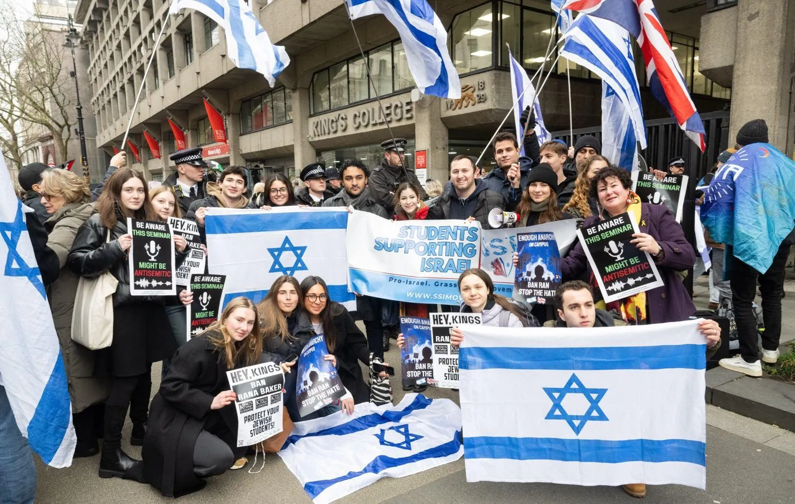 A group of students stands and kneels for a photograph outside King’s College London behind a banner reading Students Supporting Israel, holding Israeli flags and printed signs.