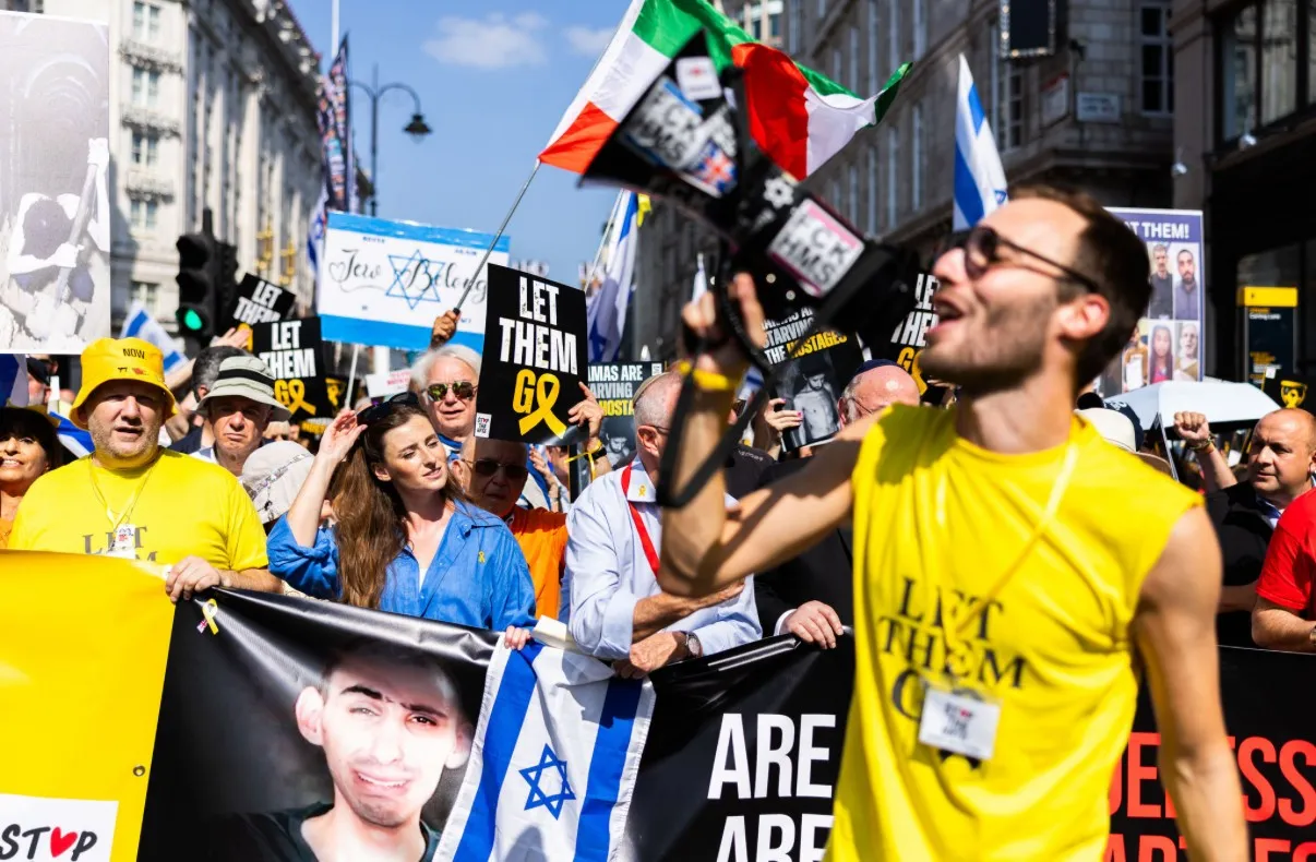 A man in a yellow Let Them Go vest shouts into a megaphone at the head of a march, hundreds behind him carrying Let Them Go placards, Israeli flags and printed hostage posters in a sunlit London street.