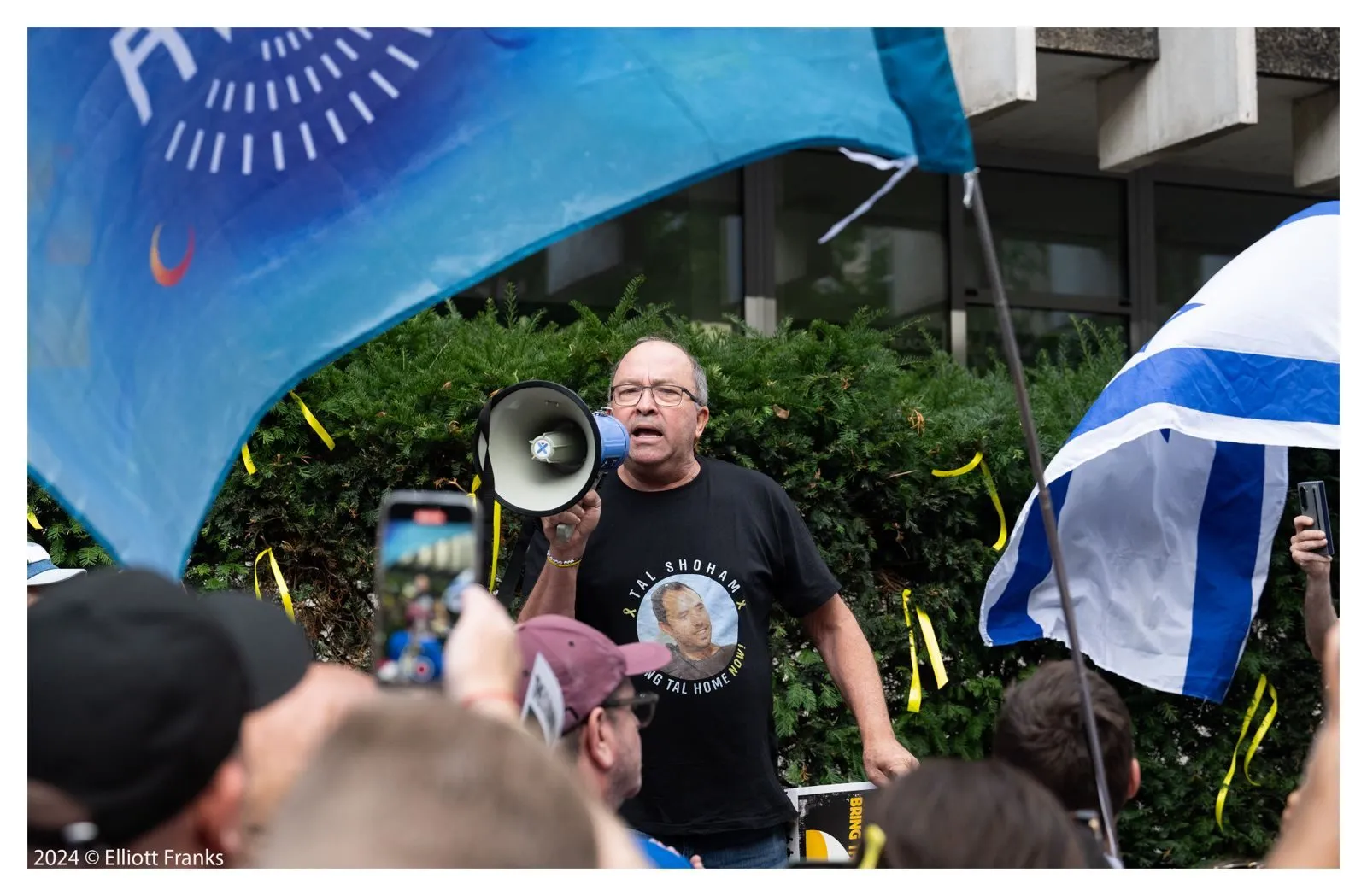 A man speaks into a megaphone to a crowd, wearing a shirt printed with a hostage portrait, a large blue flag filling the frame behind him.