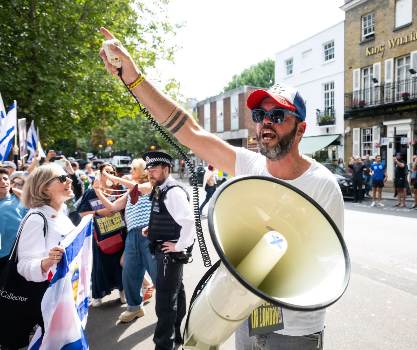 A man raises one arm and holds a megaphone in a sunlit residential street, a police officer and a line of demonstrators behind him.