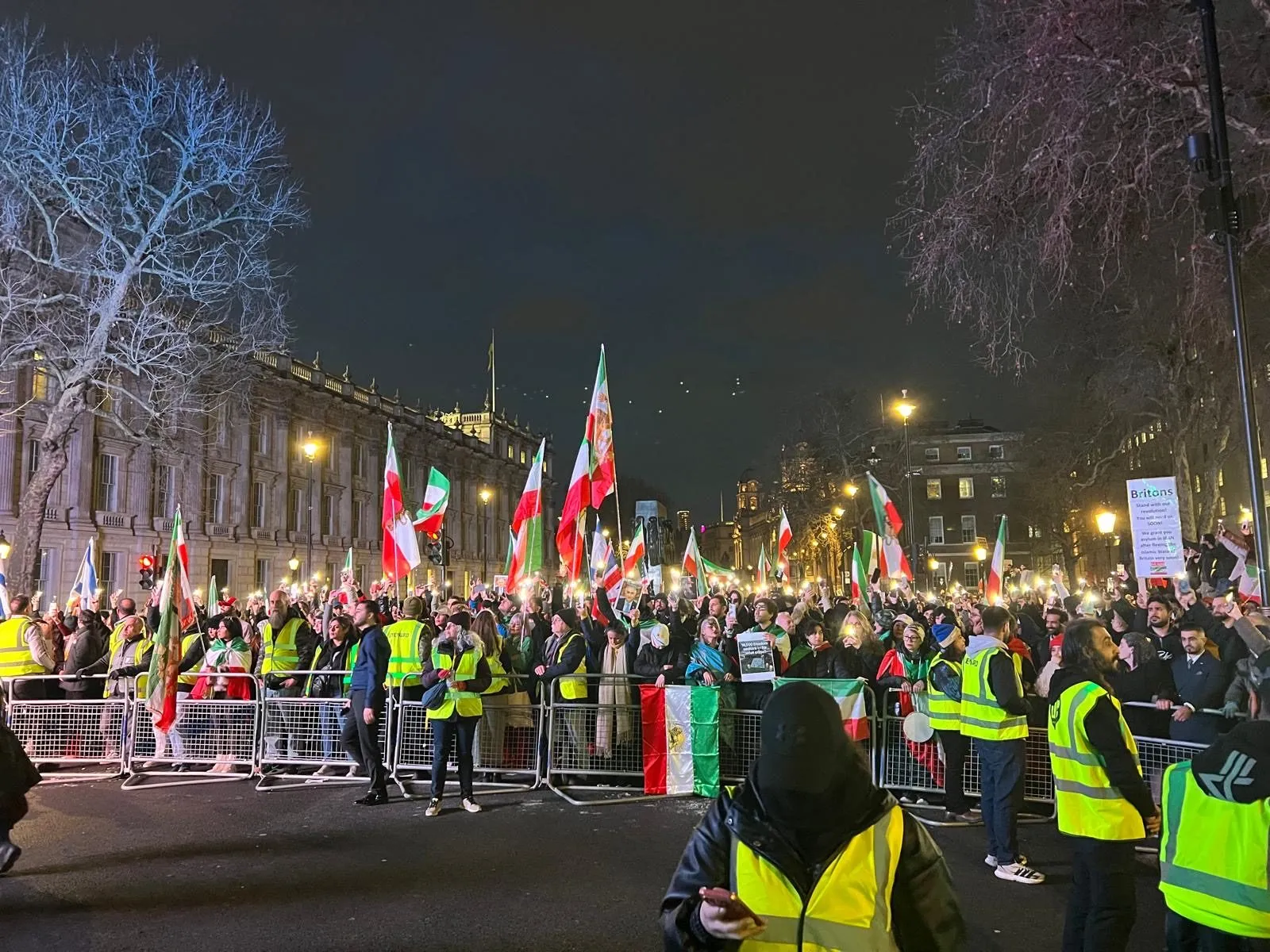 A night time crowd stands behind metal barriers on a lit London street, stewards in high visibility jackets along the front.
