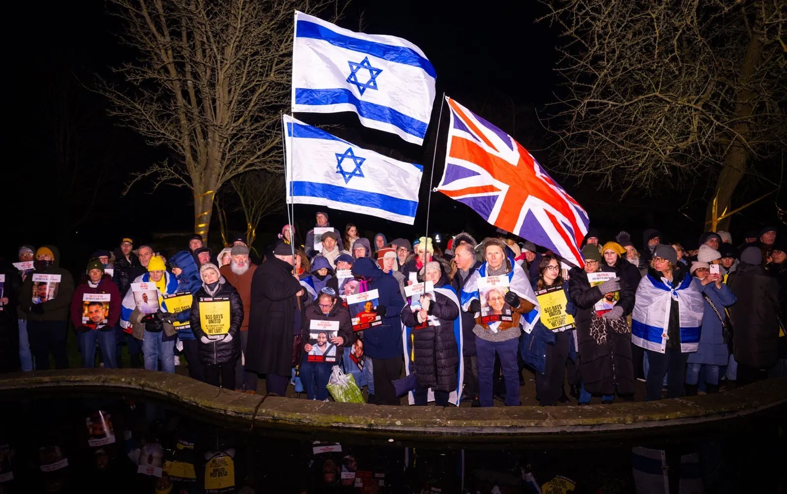 A vigil at night beside water, two Israeli flags and a Union flag lit against bare trees, the people below holding printed posters and candles.