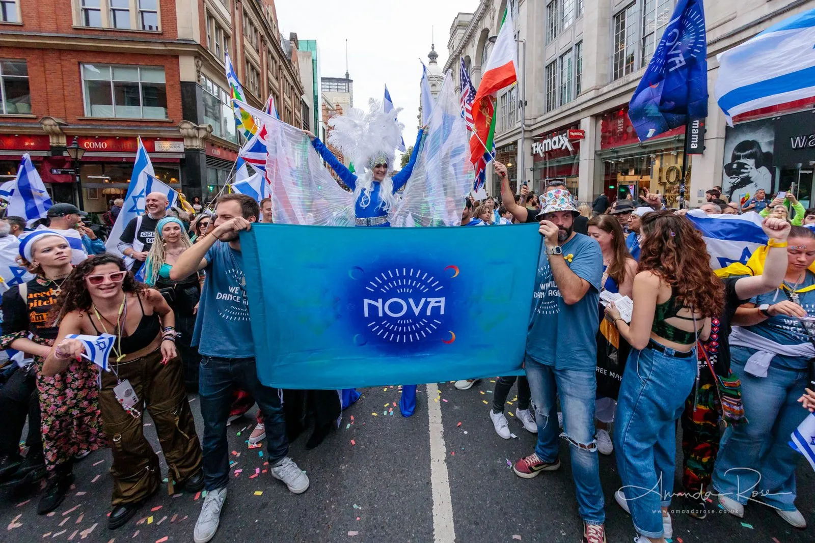 Two people carry a wide blue Nova banner down the middle of a London shopping street, confetti on the road and Israeli flags on both sides.