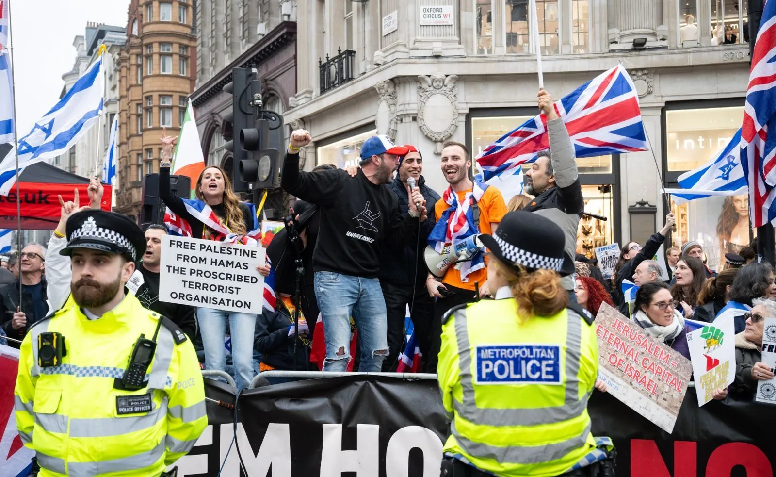 Two Metropolitan Police officers stand with their backs to the camera in front of a demonstration at Oxford Circus, a man speaking into a microphone behind them.