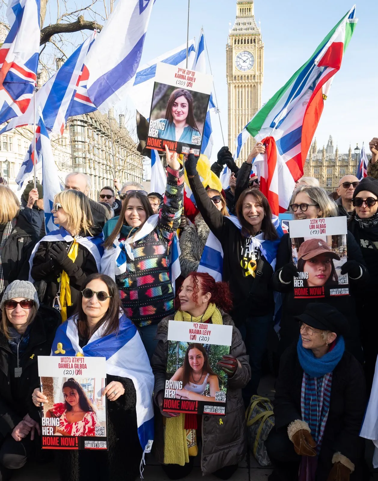 Women stand in Parliament Square holding printed placards carrying portraits of named hostages, the Elizabeth Tower and Israeli flags behind them.