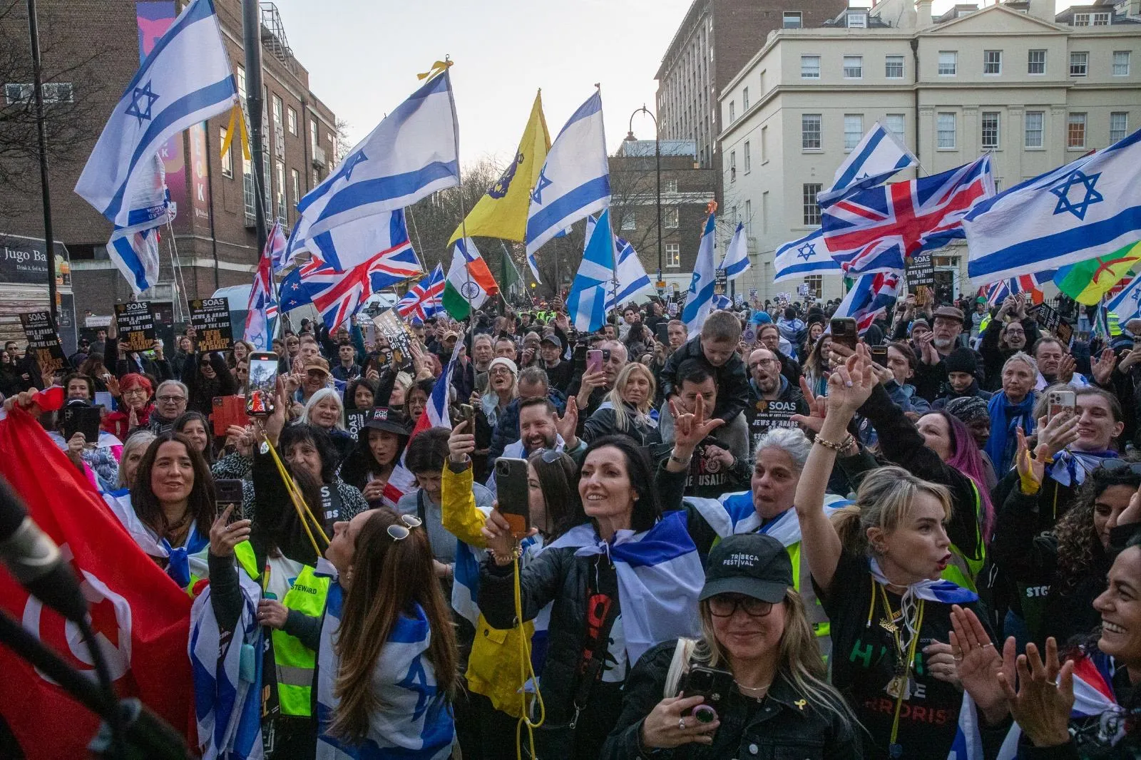 A dense crowd in a London street with Israeli and Union flags overhead, a dozen people holding phones up to film what is happening in front of them.