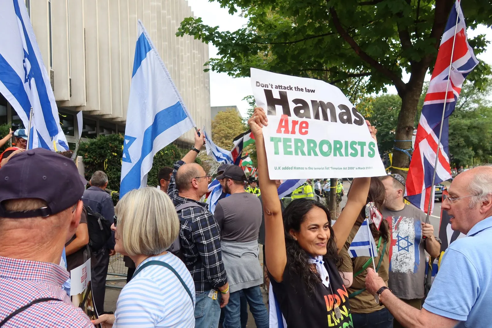 A woman holds a hand made placard reading Hamas Are Terrorists above her head in a crowd carrying Israeli and Union flags under summer trees.