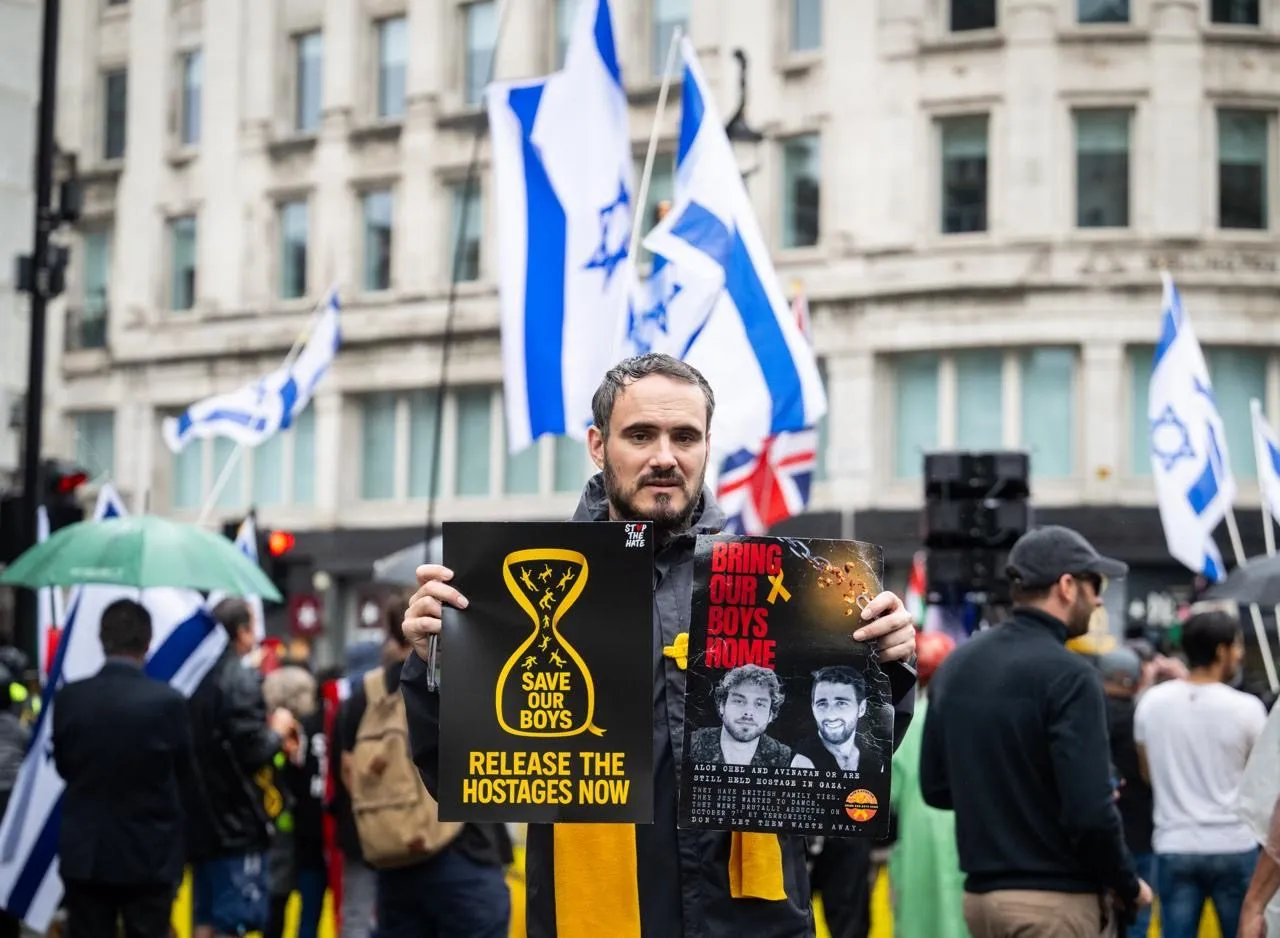A man stands in the road holding a placard reading Release The Hostages Now, Israeli flags and a stone office building behind him.