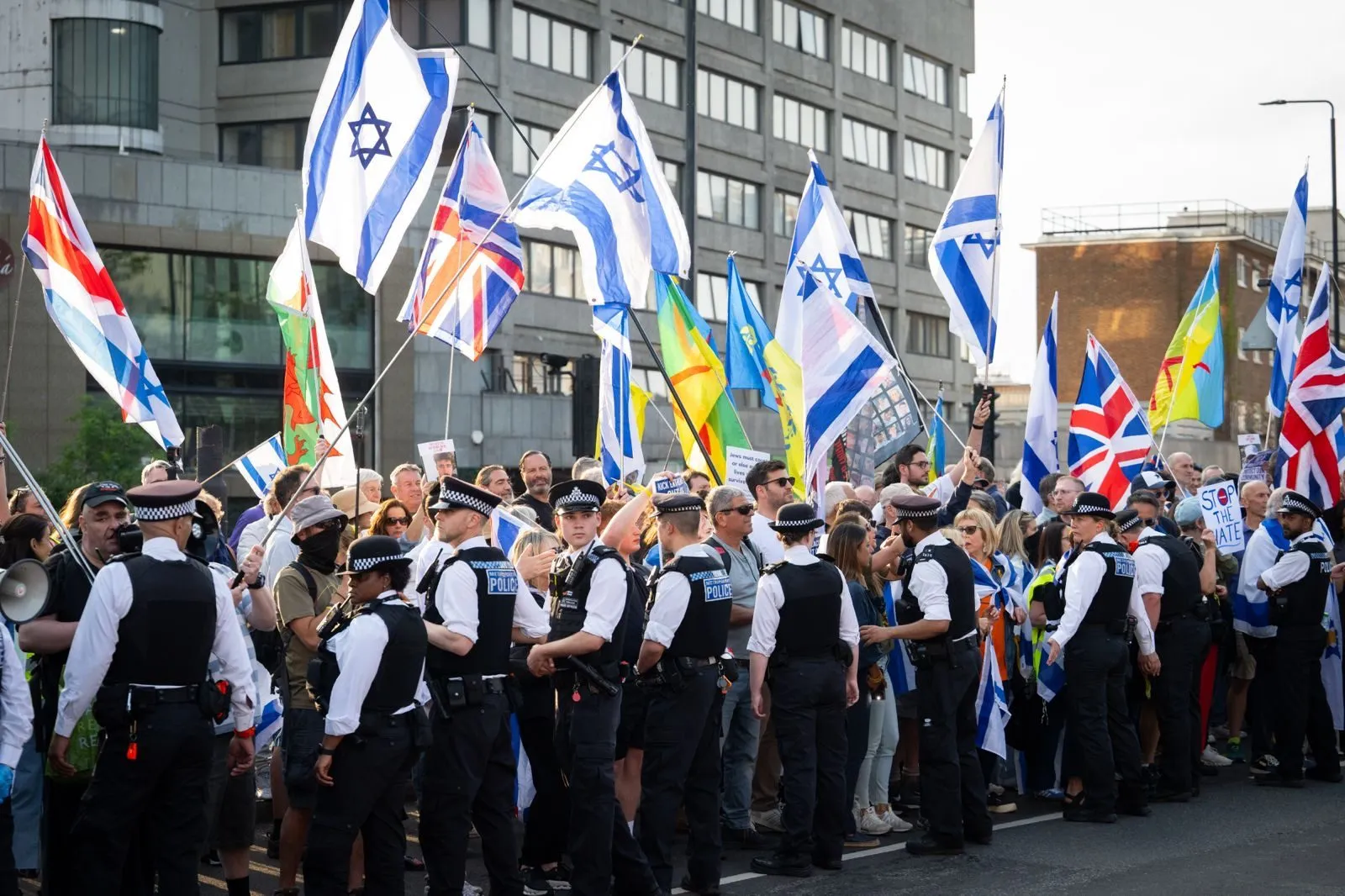 A line of police officers stands across a road in front of a crowd holding Israeli and Union flags on a summer evening.