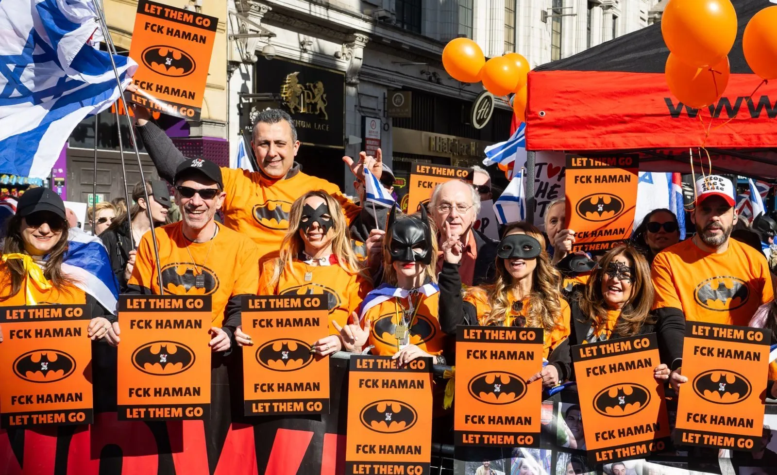A group in orange shirts and masks holds identical orange placards reading Let Them Go, in a London street with orange balloons above.