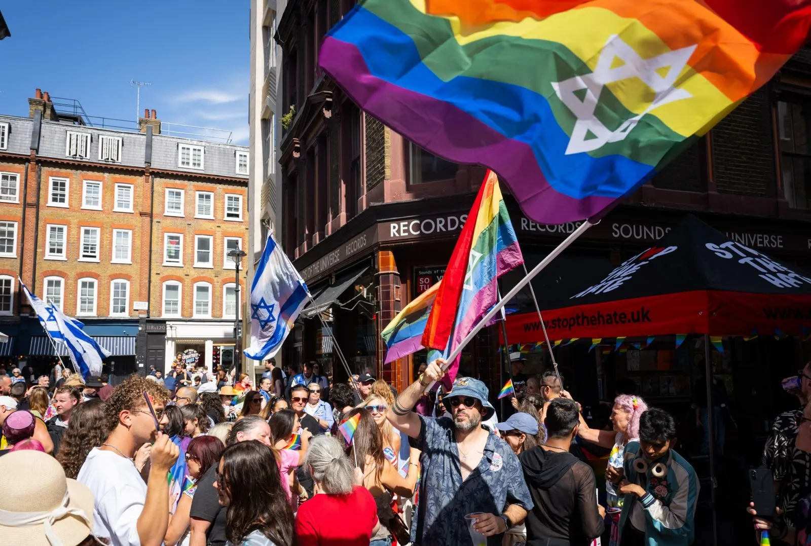 A large rainbow flag bearing a Star of David is waved above a crowded Soho street.