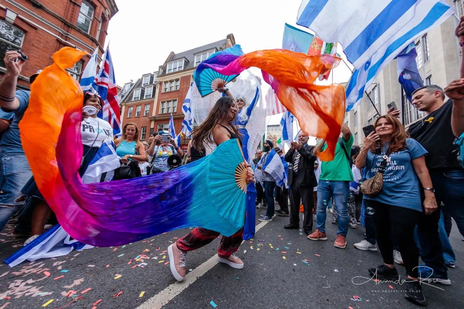 A dancer sweeps two large silk fans through the air in the middle of a London street parade, flags and onlookers on both sides.