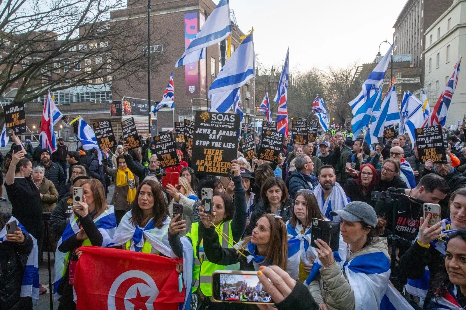 A crowd holds printed placards addressed to SOAS above their heads, many people filming on phones, Israeli flags along the street behind.