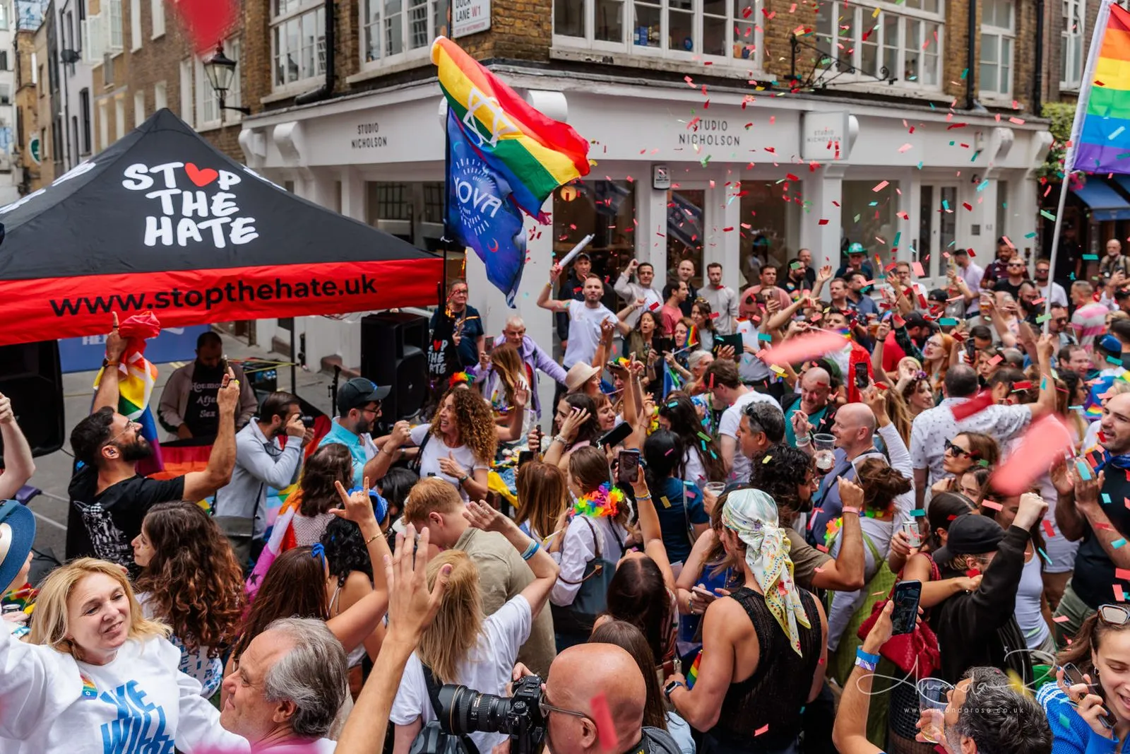 Confetti falls over a crowd with raised arms in a narrow Soho street, a black Stop The Hate marquee at the left of the frame.