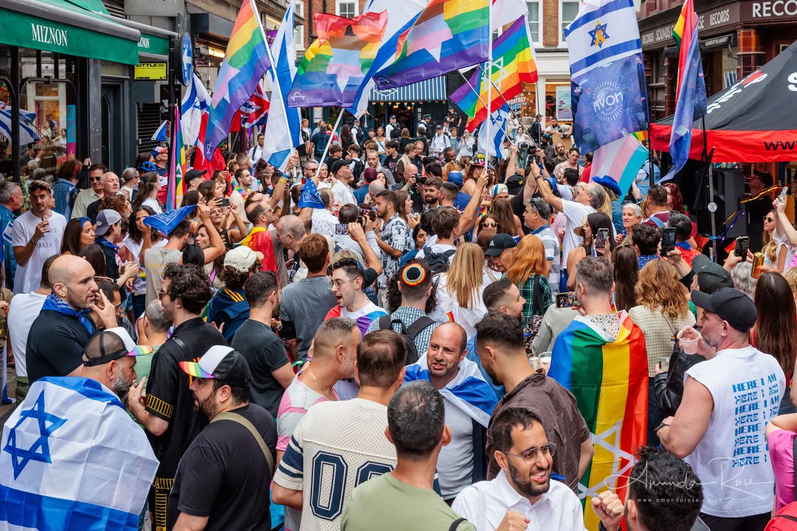 A crowd fills a Soho street between shopfronts, Israeli, rainbow and Union flags raised above it in bright sunshine.