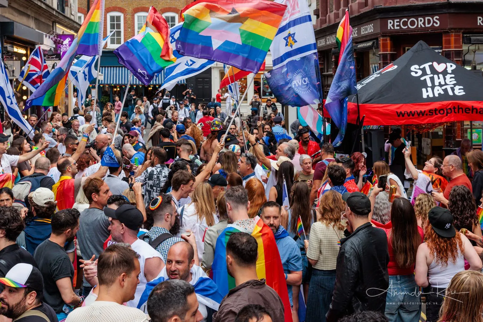 Rainbow and Israeli flags are waved above a crowd in a narrow Soho street, a black Stop The Hate marquee with its web address on the right.