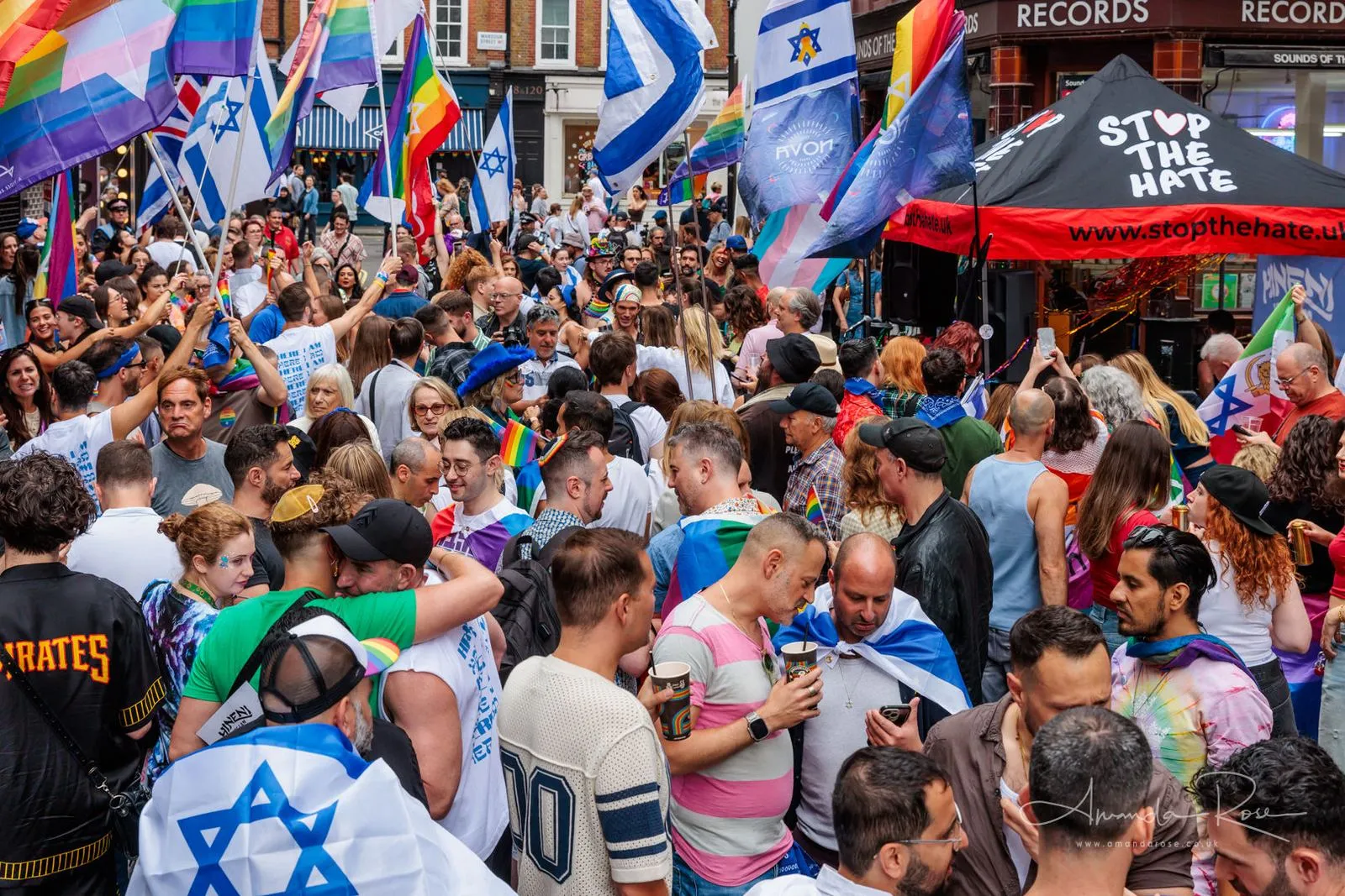A packed Soho street with rainbow and Israeli flags overhead and a black Stop The Hate marquee on the corner.