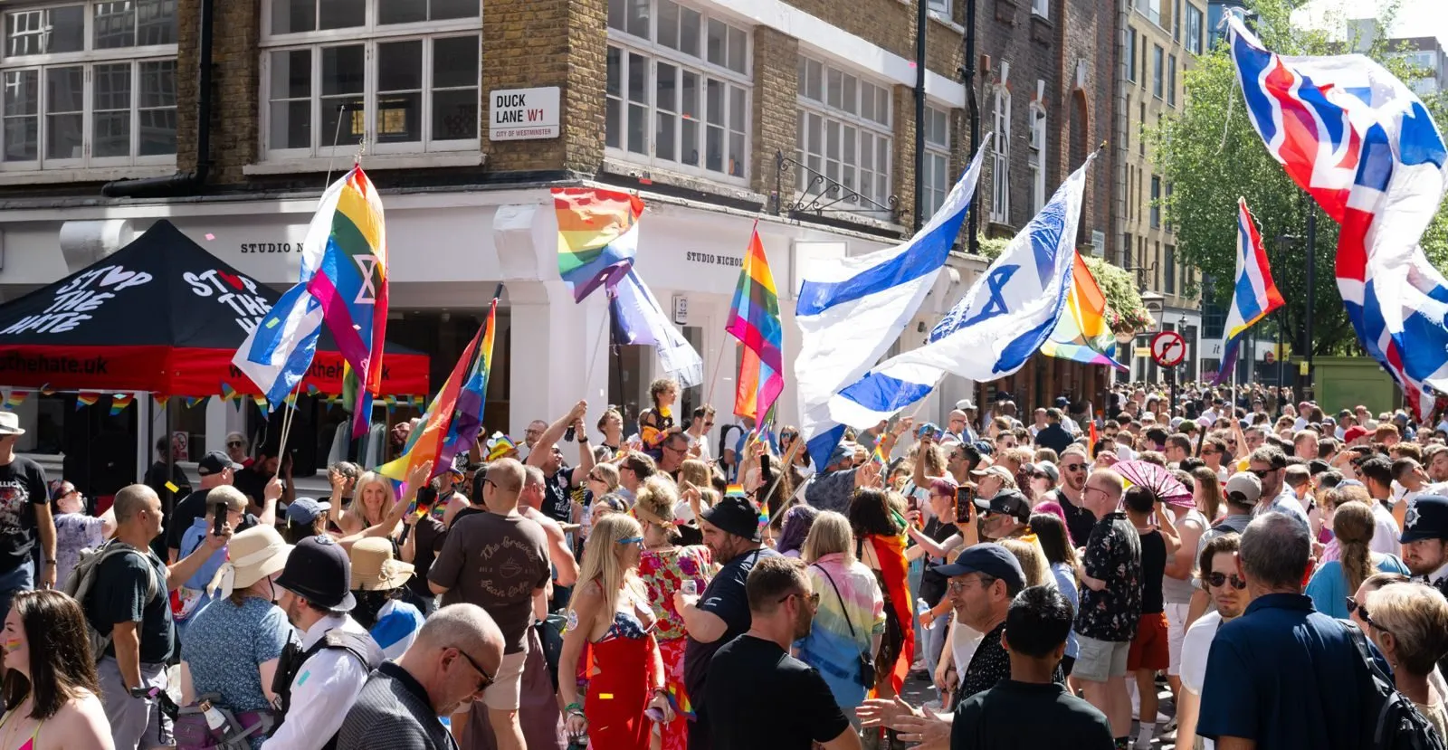 A packed street party under a Duck Lane W1 sign, rainbow and Israeli flags overhead and a black Stop The Hate marquee at the left.