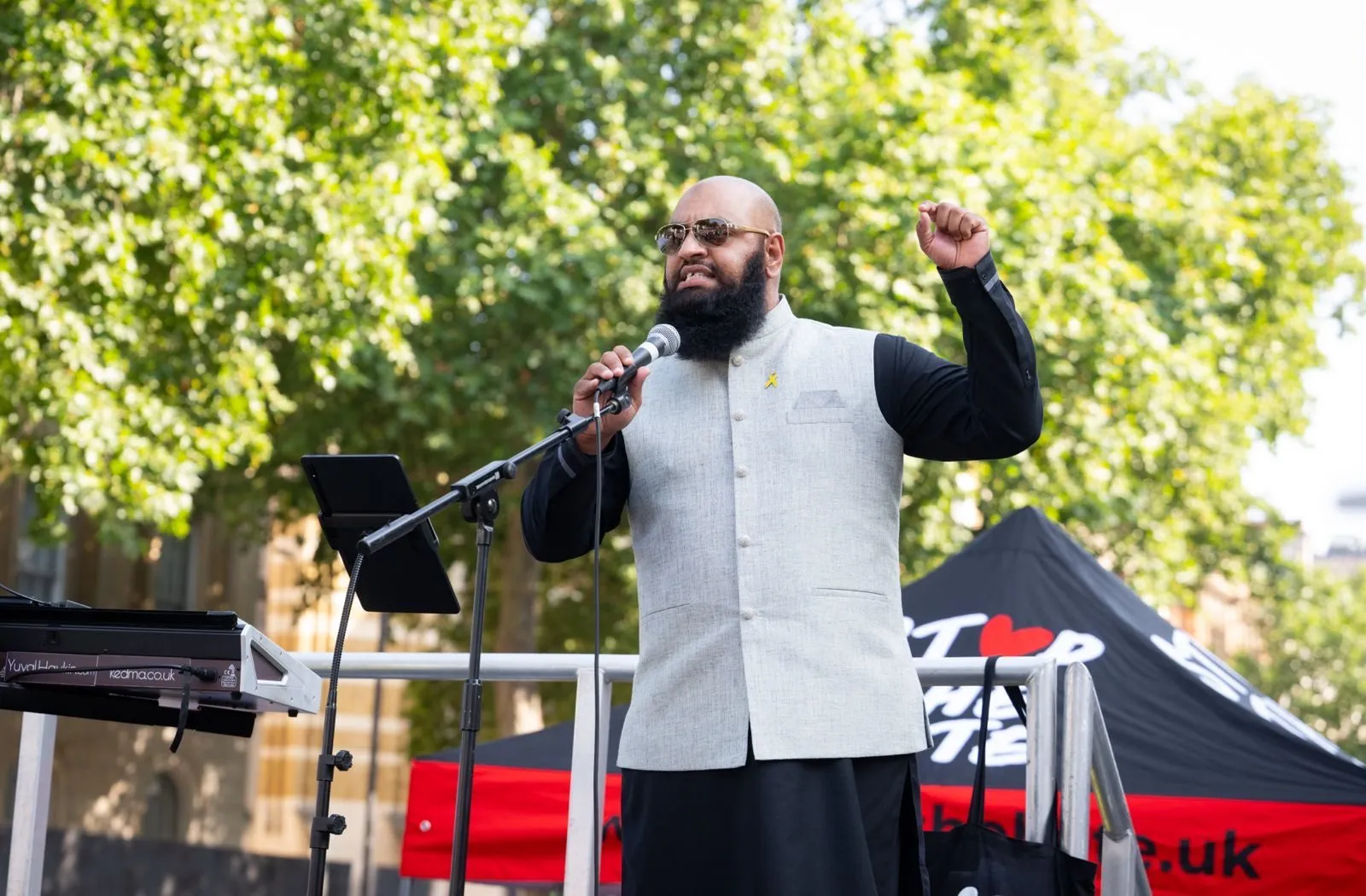 A speaker at a microphone raises a fist on an outdoor stage, a black Stop The Hate marquee and a keyboard behind him under summer trees.