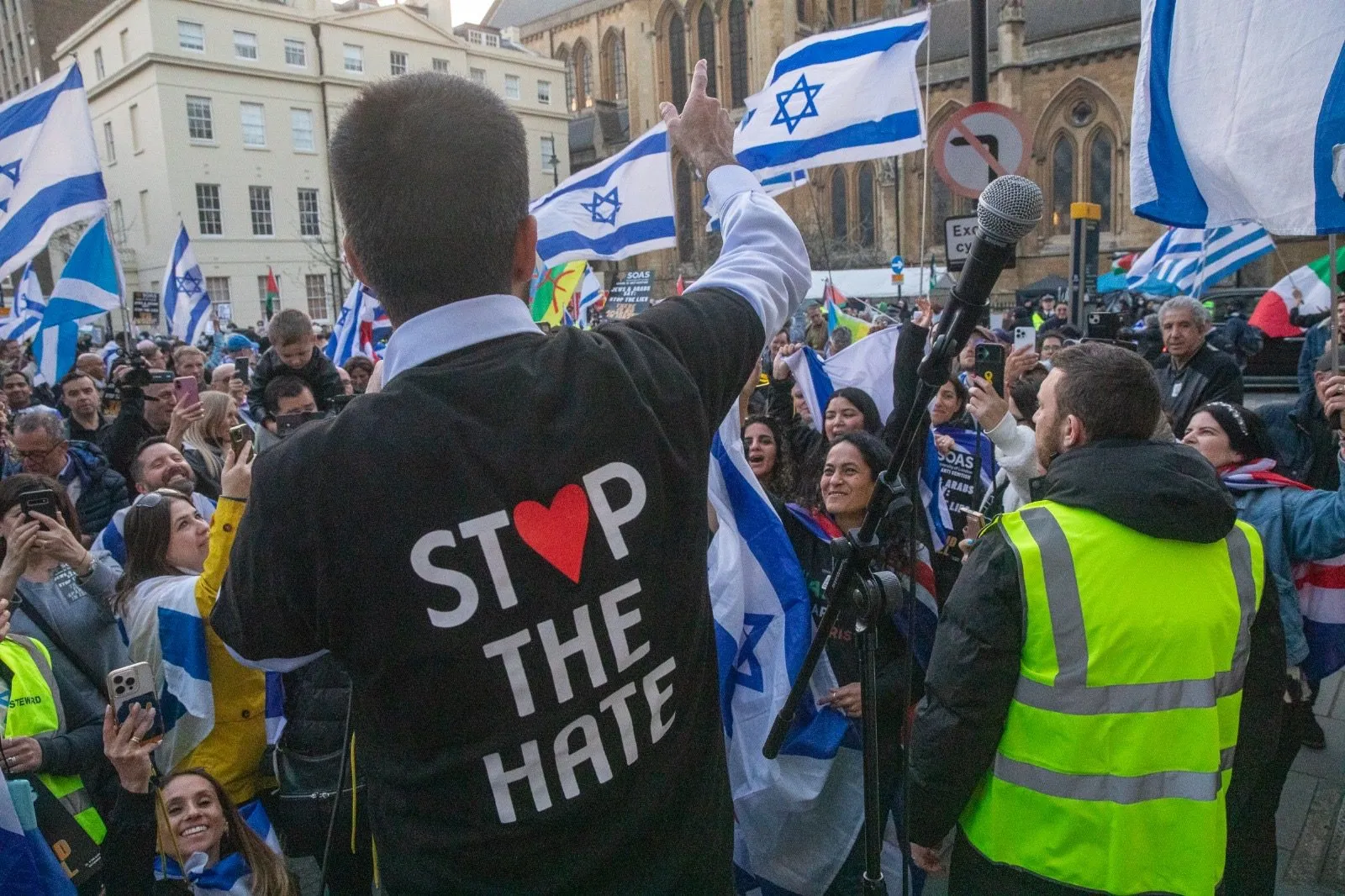 Seen from behind, a speaker in a jacket printed Stop The Hate raises an arm to a crowd of several hundred holding Israeli flags in a London street.