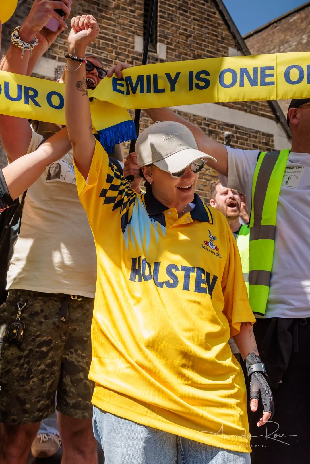 A woman in a yellow football shirt raises one fist beneath a yellow scarf reading Emily is one of our own, held up across the frame by other hands.