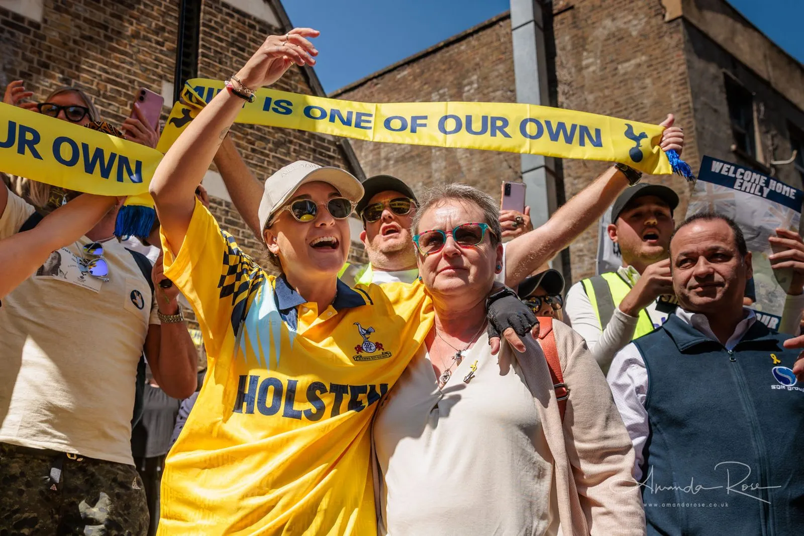 Two women hold a yellow football scarf reading Emily is one of our own above their heads in bright sunshine outside a brick stadium wall.