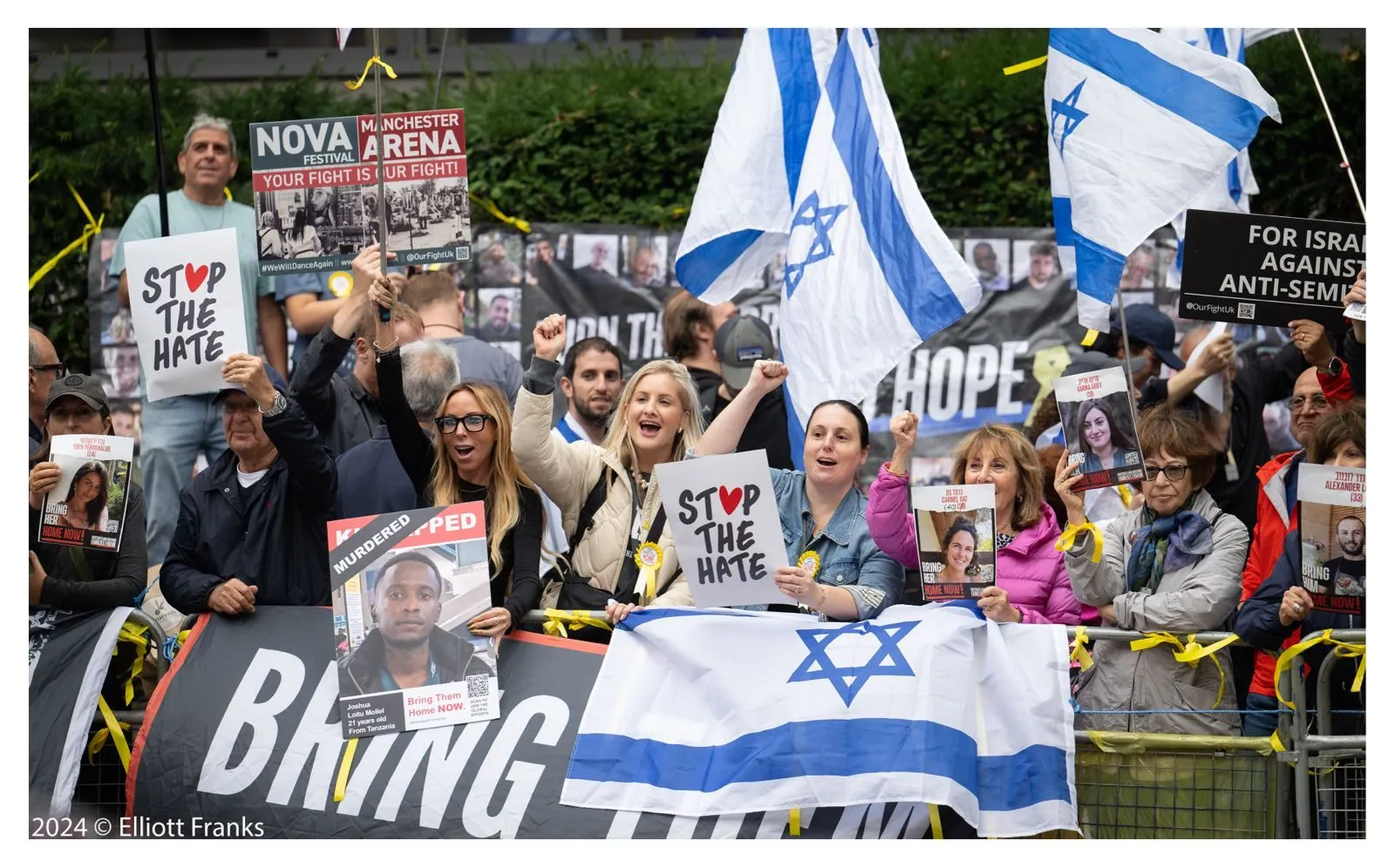 People behind a barrier hold small Stop The Hate signs and raise their fists, printed hostage posters and Israeli flags around them.