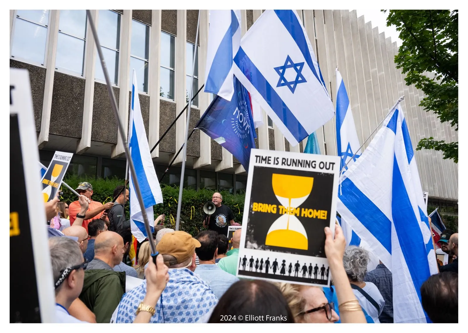 A placard reading Time Is Running Out with an hourglass is held above a crowd of Israeli flags outside a concrete office building.