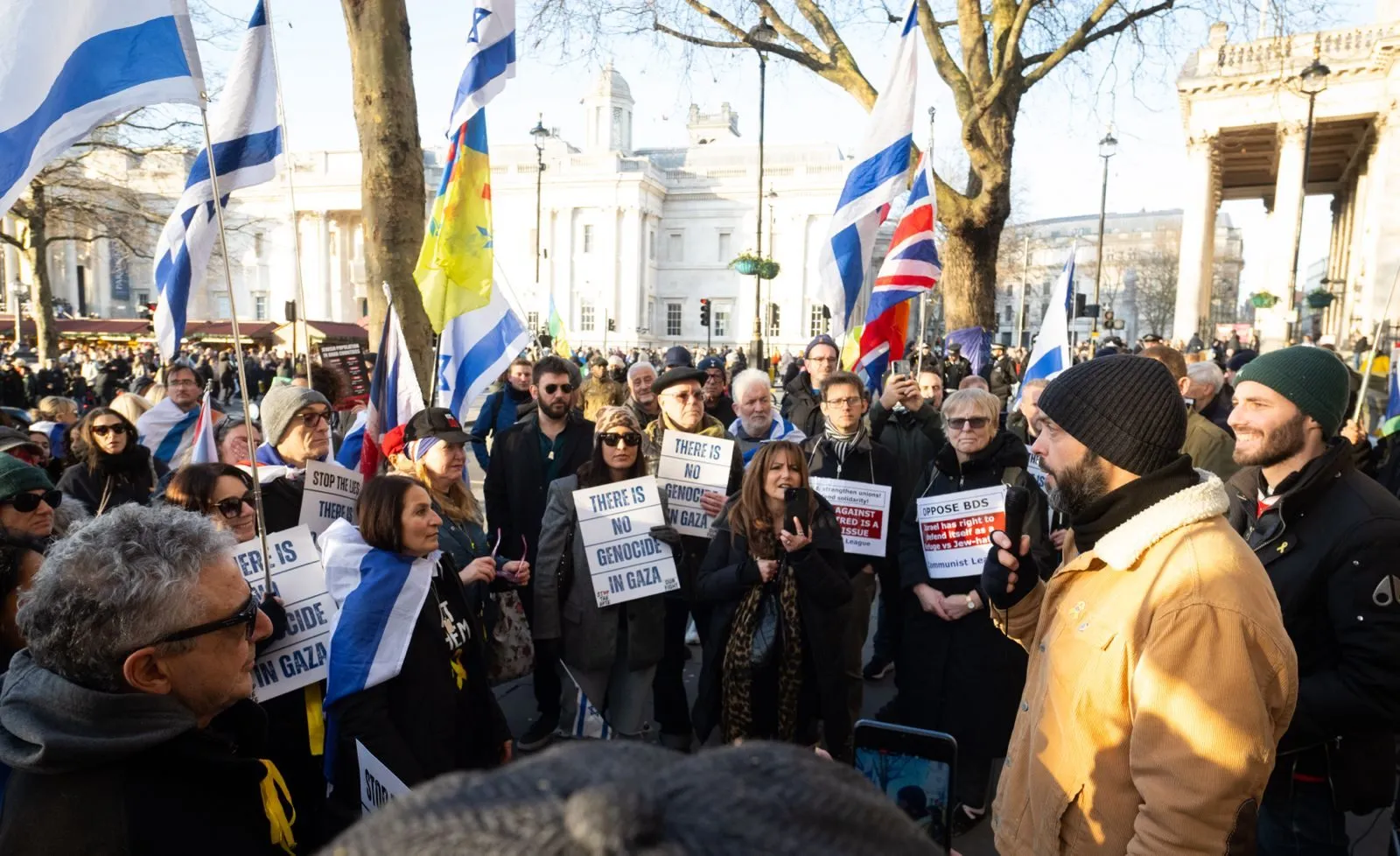 A man in an orange coat stands facing a group holding printed placards in Trafalgar Square, the two sides in conversation across a short gap.