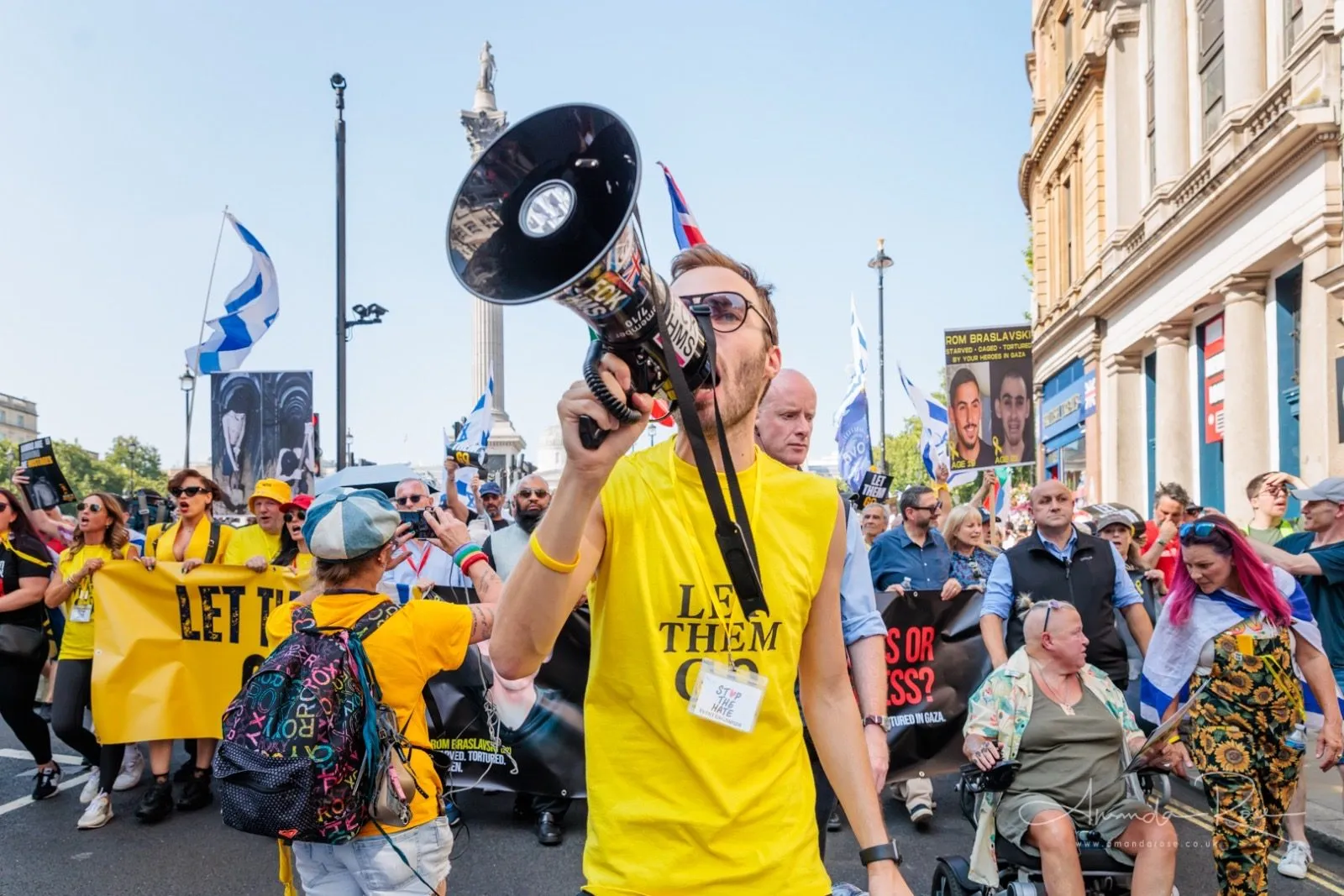 A man in a Let Them Go shirt walks backwards at the head of a march holding a megaphone up, Nelson’s Column and a yellow banner behind him.