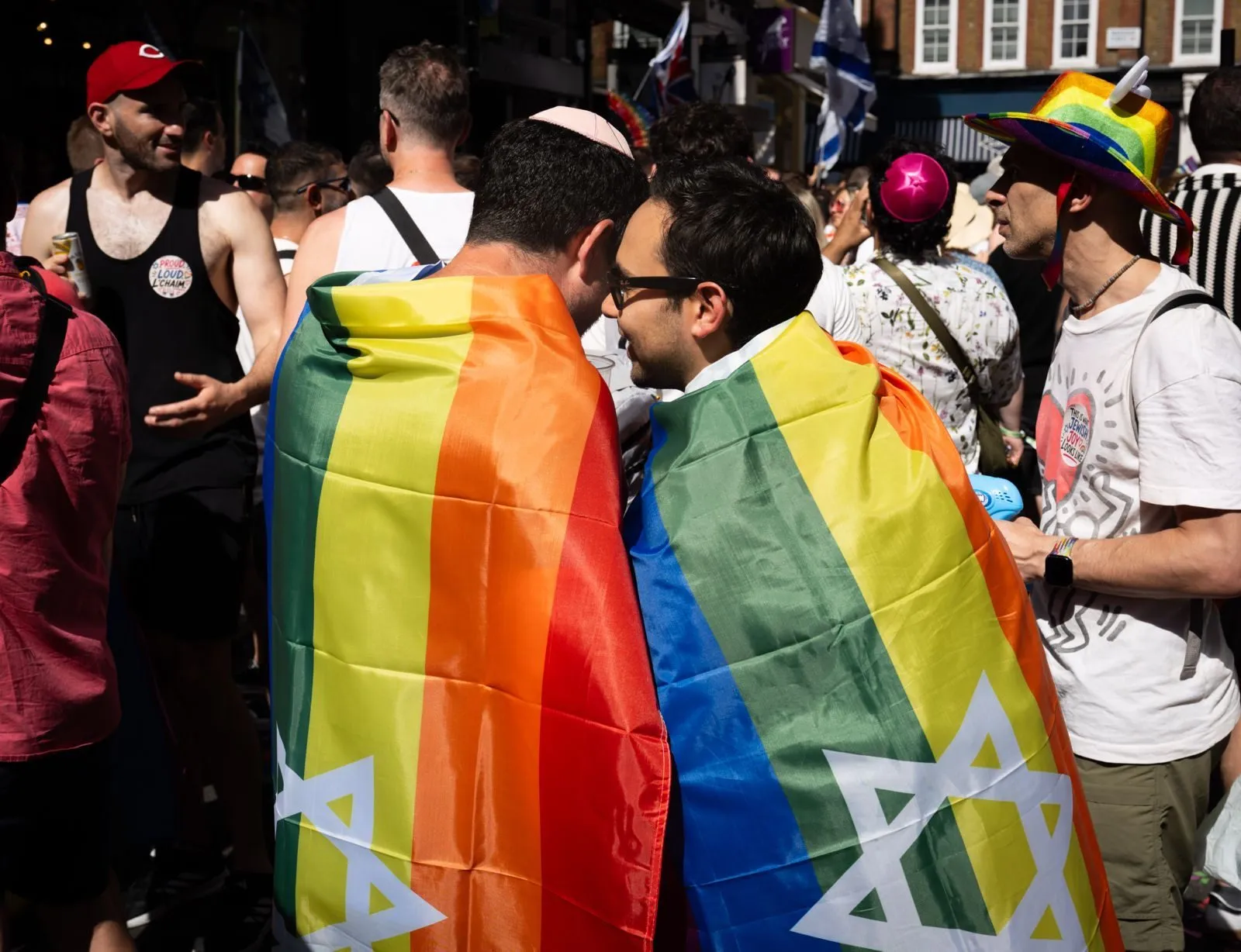 Two men wrapped in rainbow flags bearing a Star of David stand face to face talking in a crowded sunlit street.
