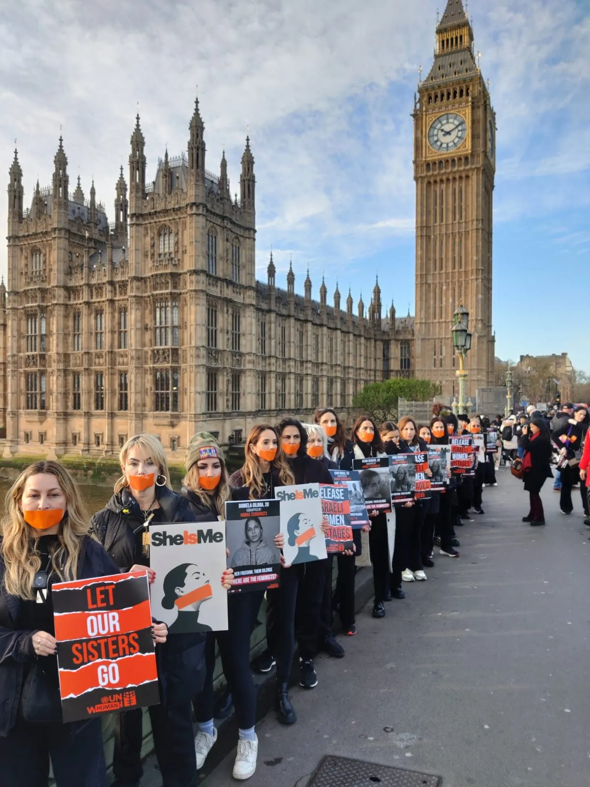 A long single file line of women in orange face masks stretches along Westminster Bridge past the Palace of Westminster, each holding a printed placard.