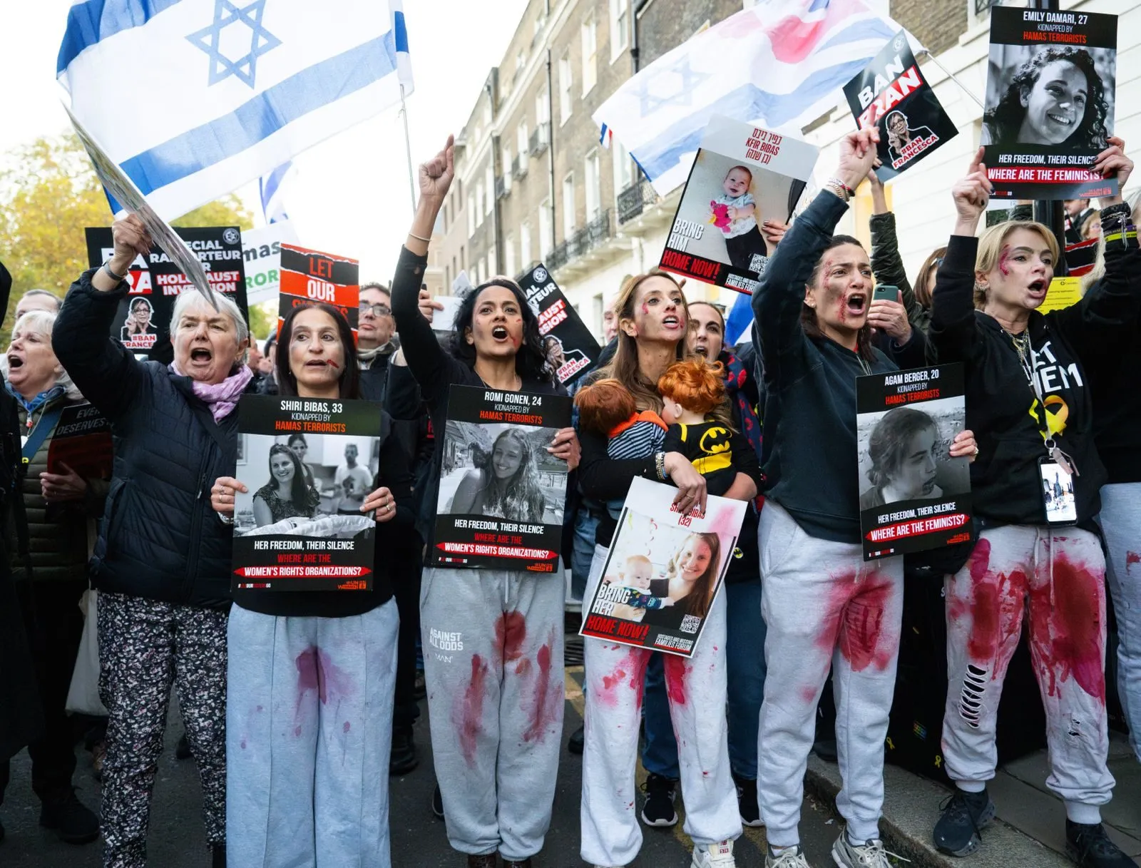 A line of women in white clothing marked with red paint hold placards asking where the feminists are, printed portraits of named hostages in their hands.
