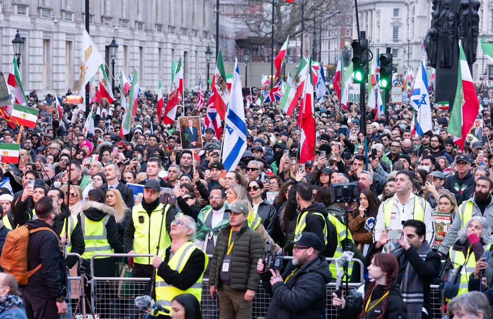 A large crowd fills Whitehall behind barriers, stewards in high visibility jackets spaced along the front of the line.